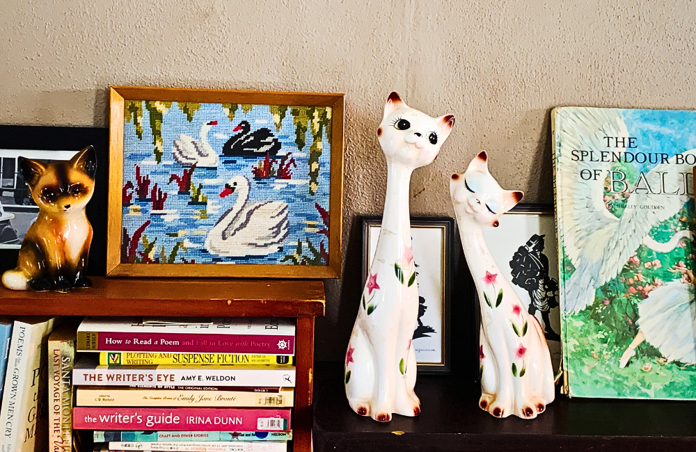 A shelf in Pip's house, featuring a ballet book, two ceramic cats, a swan tapestry, a ceramic fox and various books