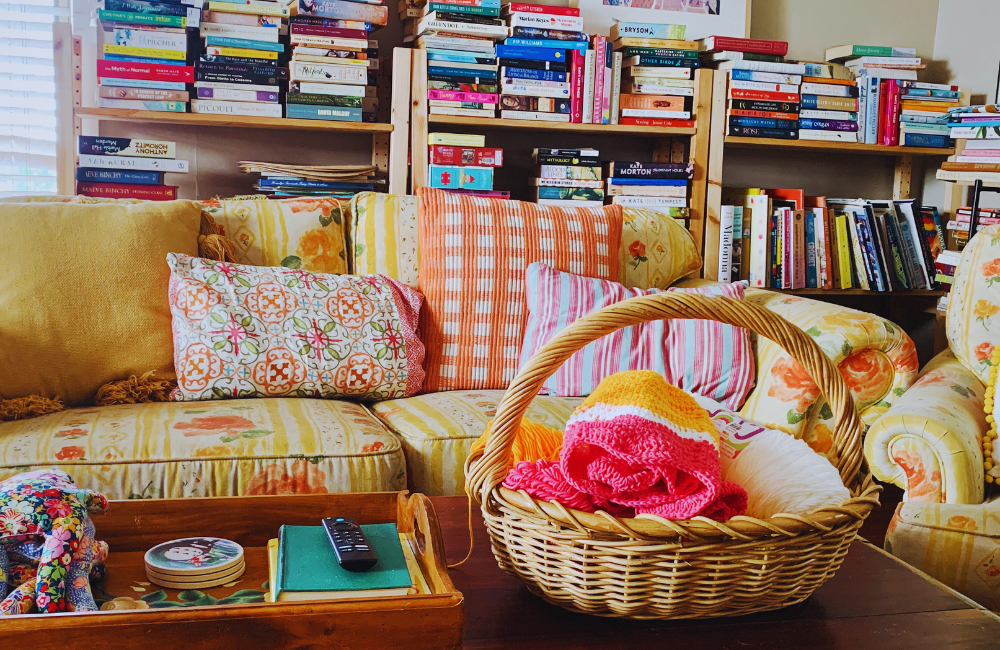 A photo of Pip's living room featuring yellow floral couch lined with cushions, behind it are some overloaded pine bookcases, in the front of the photo is a basket of crochet and a tray of books and coasters.
