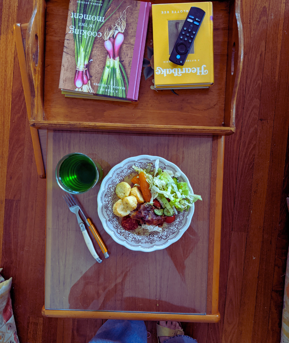 Chicken dinner on a table with a glass of green cordial and a tray of recently bought op shop books. There's also a remote control and some cutlery.