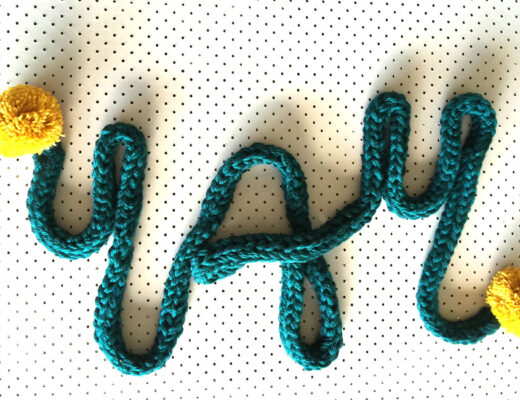 a dark green finger-knitted scarf forming the word 'YAY'. There is a yellow pompom at each end of the scarf.