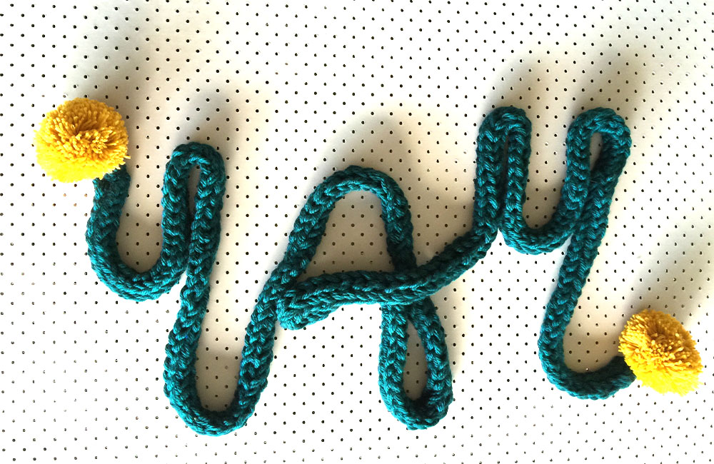 a dark green finger-knitted scarf forming the word 'YAY'. There is a yellow pompom at each end of the scarf.