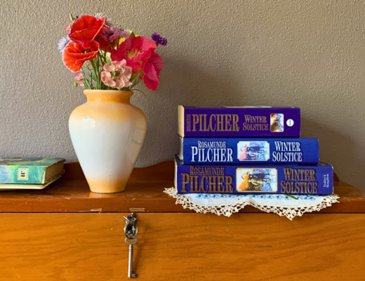 A stack of copies of Winter Solstice by Rosamunde Pilcher beside a vase of cut flowers