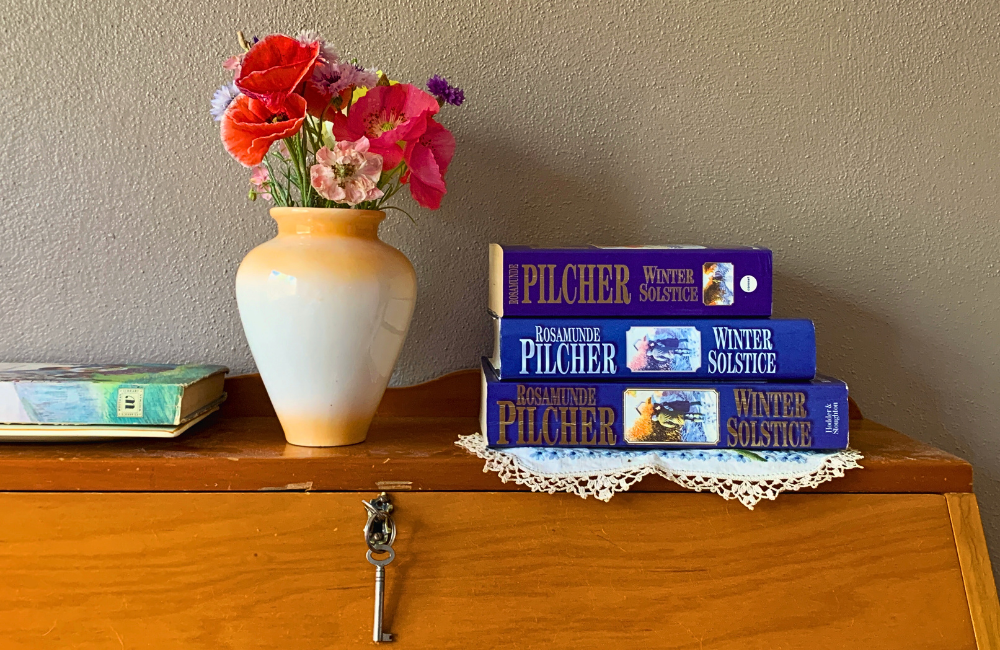 A stack of copies of Winter Solstice by Rosamunde Pilcher beside a vase of cut flowers