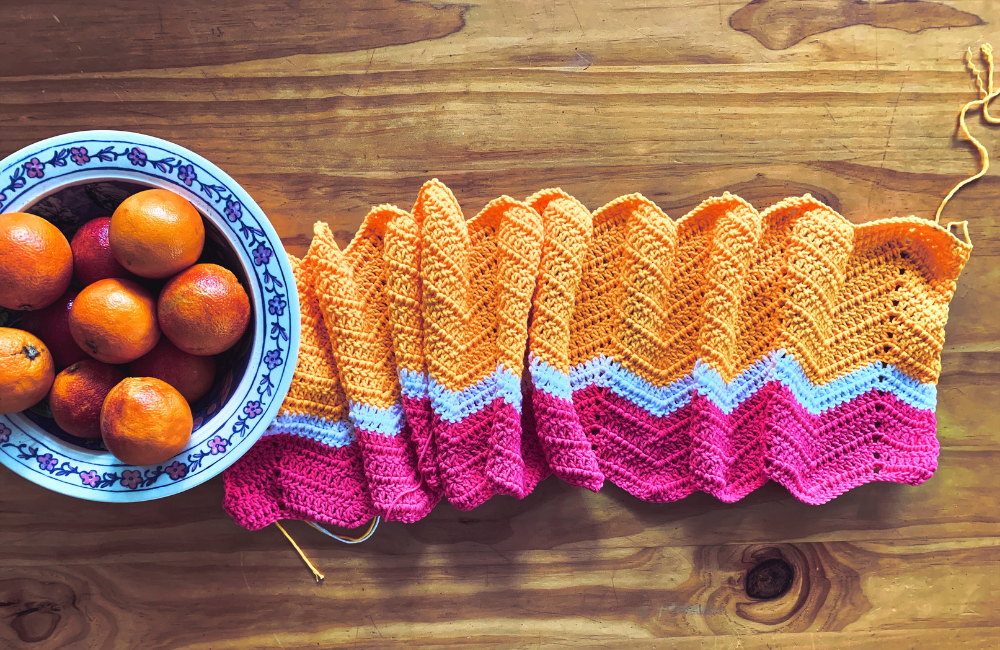 A yellow, pink and white ripple blanket on a tabletop alongside a bowl of blood oranges
